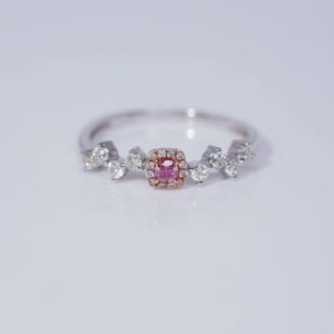 Könnte beinhalten: Ein zarter Silberring mit einem rosafarbenen Diamanten in der Mitte, der von kleineren Diamanten umgeben ist. Der Ring hat ein einzigartiges Design mit einer Diamantenansammlung auf jeder Seite des Mittelstücks.