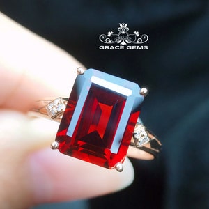 May include: A red gemstone ring with a large emerald-cut center stone and smaller diamond accents. The ring is set in a gold band.