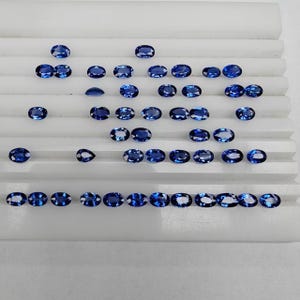 Può includere: Una collezione di numerosi zaffiri blu sfaccettati e sciolti in varie forme ovali e a goccia. Le gemme presentano tonalità blu profonde e vibranti e sono disposte in file su un vassoio espositore bianco con scanalature orizzontali.