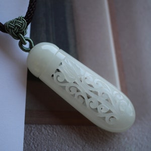 May include: A white jade pendant with intricate carved floral designs. The pendant is attached to a green cord.