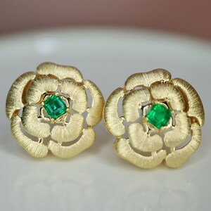 May include: A pair of gold-colored flower-shaped stud earrings. Each earring features a square-cut, vibrant green gemstone at the center. The earrings have a textured, matte finish, and the floral design is detailed with multiple petals.