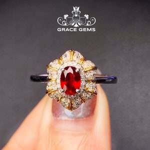 May include: A red ruby gemstone ring with a halo of smaller diamonds set in a gold band. The ring is surrounded by a larger halo of diamonds.