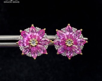 May include: A pair of flower-shaped stud earrings. Each earring features a central diamond surrounded by pink marquise-cut gemstones, with a halo of smaller white gemstones. The earrings have gold accents and are set against a black background.