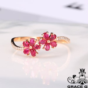 Può includere: Un anello d'oro con due motivi floreali in rubino. I fiori sono realizzati con piccole pietre di rubino rosso e sono incastonati in una fascia d'oro. La fascia è decorata con piccoli diamanti bianchi.