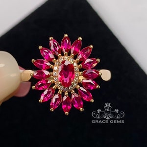 May include: A gold ring featuring a flower-like design with a central oval ruby surrounded by smaller, marquise-cut rubies. The center is encircled by small diamonds. The ring is set against a black background.