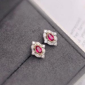 May include: A pair of ruby and diamond earrings. Each earring features a red oval ruby surrounded by a halo of small diamonds, set in a square-shaped frame.