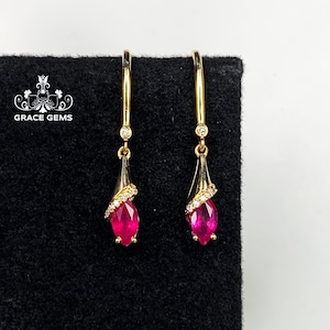 May include: A pair of gold dangle earrings featuring a red ruby gemstone and small clear diamonds. The earrings are teardrop shaped.