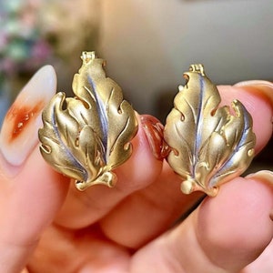 Könnte beinhalten: Ein Paar goldene Ohrringe in Blattform mit einem strukturierten Design. Die Ohrringe bestehen aus zwei Blättern, die am Stiel miteinander verbunden sind.