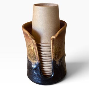 May include: A ceramic cup holder with a tall, cylindrical shape and a U-shaped cutout on the front. The exterior features a gradient glaze transitioning from a dark, glossy black at the base to a textured, mottled brown at the top rim.
