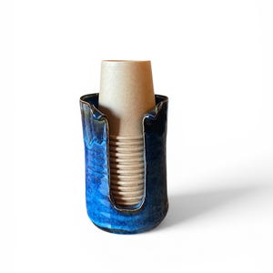 May include: A ceramic cup holder with a glossy blue glaze. The holder has a U-shaped opening and contains a brown paper cup. The holder's design features a dark blue rim and a lighter blue interior.