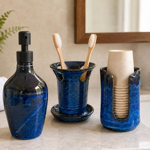 May include: A bathroom accessory set featuring a dark blue glazed ceramic soap dispenser with a black pump, a matching toothbrush holder with two bamboo toothbrushes, and a cup holder containing a stack of light beige disposable paper cups.