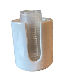 May include: A white plastic cup dispenser with a curved design. The dispenser holds a stack of clear plastic cups. The dispenser is designed to be mounted on a wall or placed on a counter.