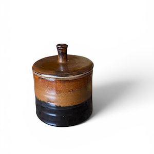 May include: A cylindrical ceramic canister with a lid featuring a small knob handle. The container has a two-tone glaze, with a glossy dark brown or black finish on the lower half and a lighter, warm tan or ochre glaze on the upper portion.