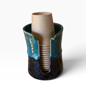 May include: A cylindrical ceramic cup holder featuring a two-tone glaze with a glossy teal upper section and a dark, textured black base. The holder has a vertical cutout opening in the front, containing a stack of light brown paper cups.