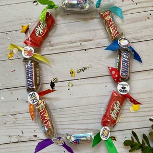 May include: A candy lei arranged in a circular shape, featuring various wrapped candy bars like Kit Kat, Snickers, and Twix. Each candy is secured with colorful ribbons and decorative stickers, with confetti and the word "Congrats" scattered around.