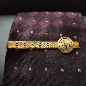May include: Gold tie clip with a patterned design and a round, engraved 'RD' charm. The tie clip is on a dark purple and white patterned tie.