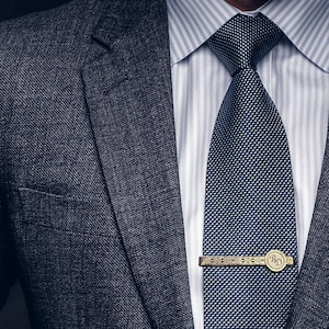 May include: A dark gray suit jacket with a white dress shirt and a dark blue patterned tie. A gold tie bar with the initials "R.D." is visible.
