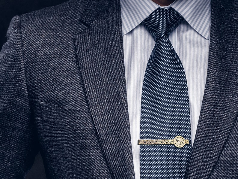 May include: A dark gray suit jacket with a white dress shirt and a dark blue patterned tie. A gold tie bar with the initials "R.D." is visible.