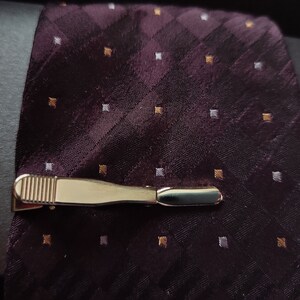 May include: A gold tie clip with a textured surface on a dark purple patterned tie with white and gold accents.