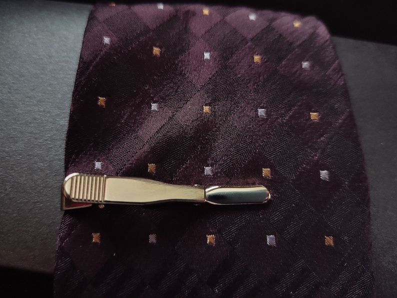 May include: A gold tie clip with a textured surface on a dark purple patterned tie with white and gold accents.