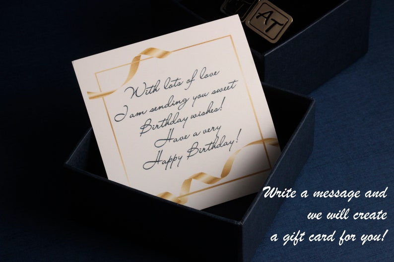 Puede incluir: Una caja de regalo azul marino con una tarjeta crema en su interior. La tarjeta tiene detalles de cinta dorada y texto escrito a mano: &laquo;&iexcl;Con mucho amor, te env&iacute;o dulces deseos de cumplea&ntilde;os! &iexcl;Feliz cumplea&ntilde;os!&raquo;. Tambi&eacute;n se ve un dije met&aacute;lico con las letras &laquo;AT&raquo;.