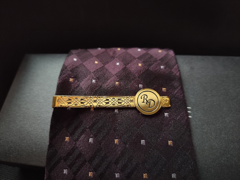 May include: Gold tie clip with a patterned design and a round, engraved 'RD' charm. The tie clip is on a dark purple and white patterned tie.