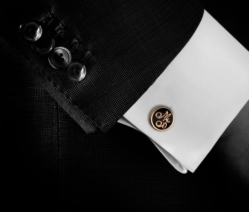 Puede incluir: Primer plano de una chaqueta de traje negra con un pu&ntilde;o de camisa blanco. Un gemelo negro con borde dorado con las iniciales "M&S" en cursiva es visible. La chaqueta tiene cuatro botones visibles.