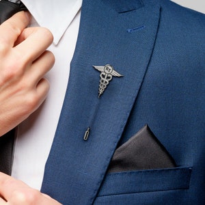 Pode incluir: Alfinete de lapela prateado com o símbolo médico do caduceu, com as iniciais "RT" num círculo. O alfinete está preso a um casaco azul. Um lenço de bolso preto é visível.