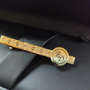 May include: A gold tie bar with an intricate design and a circular monogram with the initials "RD".