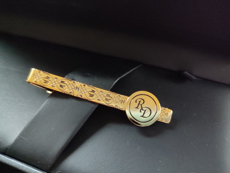 May include: A gold tie bar with an intricate design and a circular monogram with the initials "RD".