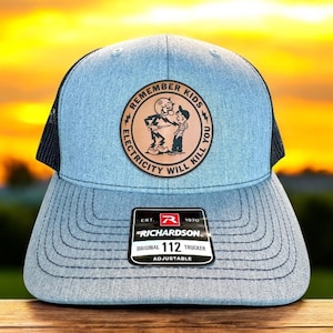 May include: A blue and black trucker hat with a brown leather patch that says "Remember Kids Electricity Will Kill You". The patch features a cartoon image of a man holding a knife and a boy. The hat is adjustable and has a Richardson 112 label.