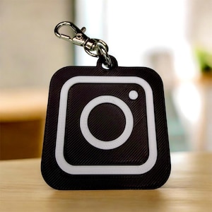 May include: Black and white Instagram logo keychain with a silver clasp. The keychain features a square design with rounded corners, a white border, and a white camera icon in the center. The background has a textured pattern.