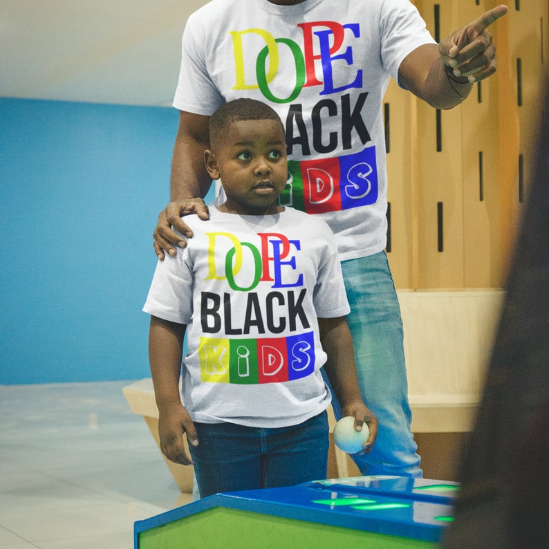 May include: Two people wearing white t-shirts with colorful text. The text on the shirts says "Dope Black Kids". The person in the front is a young boy holding a small ball.