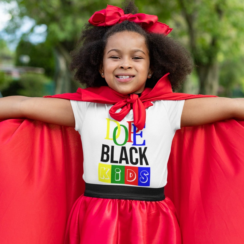 May include: A young person wearing a white t-shirt with the text "Dope Black Kids" in colorful letters. The person is wearing a red cape and a red bow in their hair. The person is smiling and looking at the camera.