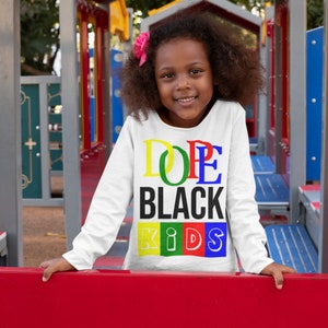 May include: A white long-sleeve shirt with the words "Dope Black Kids" printed in colorful letters. The letters are outlined in black and each letter is a different color: D is green, O is yellow, P is red, E is blue, B is black, L is yellow, A is red, C is blue, K is green, I is yellow, D is red, S is blue.