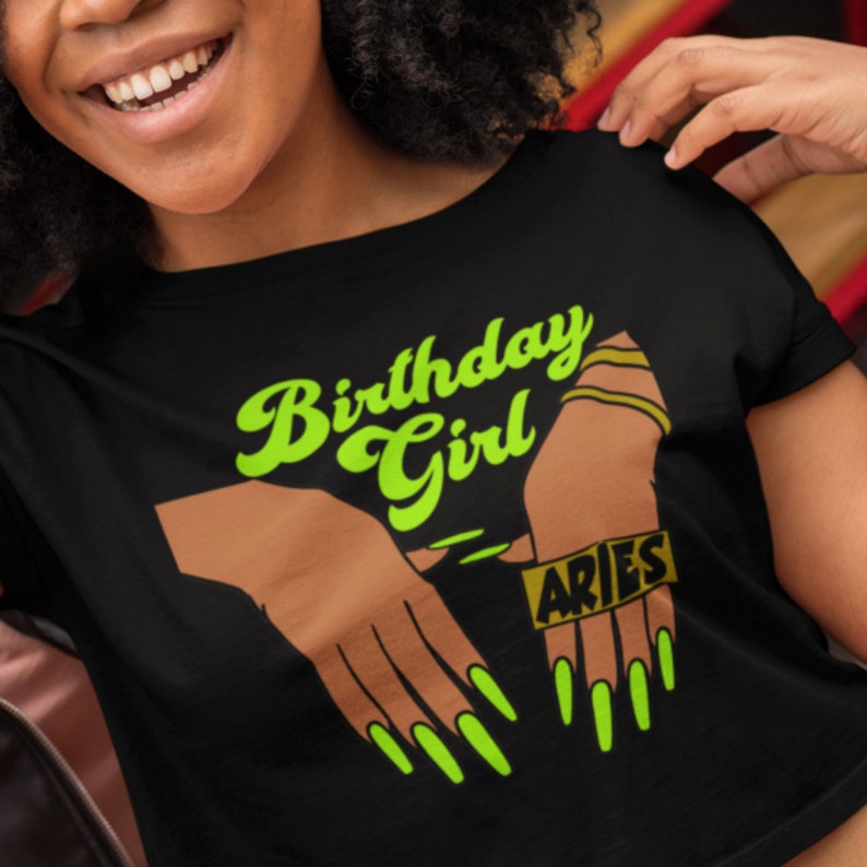 May include: Black cropped t-shirt with a green and gold graphic design. The graphic features a pair of hands with long green nails and the text "Birthday Girl" and "Aries".