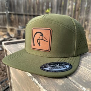 May include: A green and brown baseball cap with a duck logo on the front. The cap has a brown leather patch with a duck silhouette and the text "SNAPBACK" on the back. The cap is made of a high-quality, breathable material.
