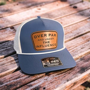May include: A blue and white trucker hat with the text "OVER PAR AND UNDER THE INFLUENCE" on a brown patch. The hat has a white rope detail and a tag that reads "WATERPROOF".