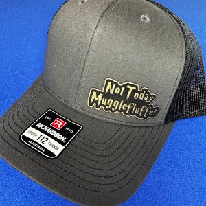 May include: A gray and black trucker hat with a gold patch that says "Not Today Mugglefluffer". The hat is adjustable and has a Richardson 112 label.