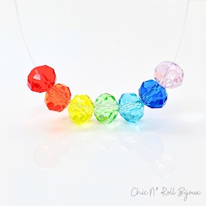 May include: A rainbow-colored necklace featuring faceted beads in red, orange, yellow, green, blue, and pink. The beads are strung on a clear, nearly invisible cord. The necklace is displayed on a white surface. The text "Chic N' Roll Bijoux" is in the bottom right corner.