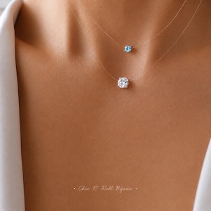 May include: A double-layered necklace featuring two clear nylon threads, each holding a single round-cut stone. The top stone is light blue, while the larger bottom stone is clear and colorless. The text Chic N Roll Bijoux is visible at the bottom.