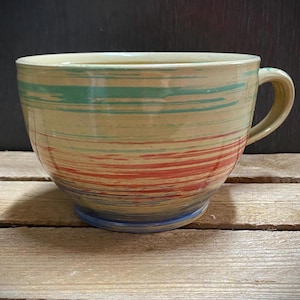 Könnte beinhalten: Ein Keramikbecher mit cremefarbener Basis und horizontalen Pinselstrichmustern in Türkis, Rot und Blau. Der Becher hat eine breite, abgerundete Schalenform, eine glatte, glänzende Oberfläche und einen einzelnen geschwungenen Henkel an der Seite.