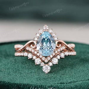 May include: A pear-shaped aquamarine engagement ring set in rose gold with a halo of smaller diamonds. The ring is on a green velvet surface.