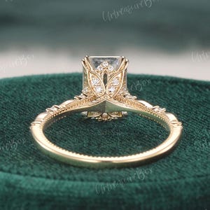 May include: A gold ring featuring a rectangular centre stone held by a decorative setting with intricate metalwork and small accent stones. The band has a beaded texture along the edges, and the gallery displays a floral-inspired metal design.