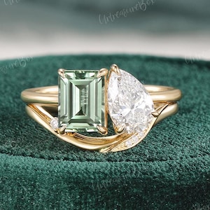 May include: A gold engagement ring featuring a rectangular green gemstone and a pear-shaped white gemstone. The ring has a delicate, curved band with small accent stones. The setting is on a green velvet surface.