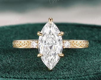May include: A marquise-cut diamond engagement ring with a gold band. The ring features a large, clear marquise-shaped diamond as the centerpiece, with smaller diamonds and an ornate design on the band. The ring is set against a dark green background.