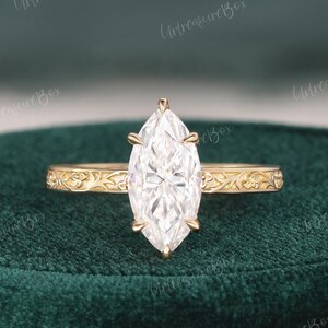 May include: A gold ring featuring a marquise-cut clear gemstone set in a six-prong mounting. The band is crafted from yellow gold and displays an intricate, embossed floral pattern that extends along the sides of the ring shank.