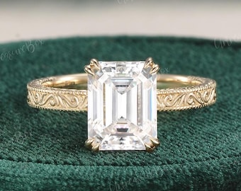 May include: A gold engagement ring with an emerald-cut diamond. The ring features an ornate, engraved band. The diamond is set with four prongs. The ring is displayed on a green velvet surface.