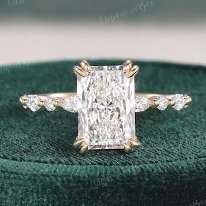 Pode incluir: Um anel de noivado de ouro com uma grande pedra central de diamante retangular e transparente. A banda é adornada com diamantes redondos menores de cada lado da pedra central.