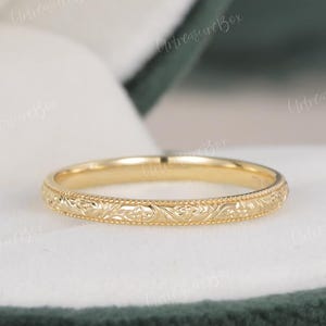 Può includere: Un sottile anello a fascia in oro giallo caratterizzato da un intricato motivo floreale inciso sulla superficie esterna, bordato da delicati dettagli a perline. Il metallo ha una finitura lucida che riflette la luce sul design in rilievo.